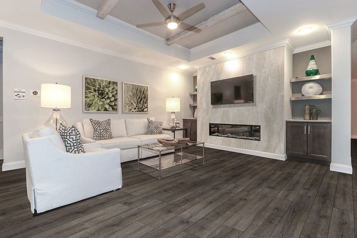Modern living room featuring MSI XL Cyrus Walnut Waves Luxury Vinyl Planks, a white sectional sofa, glass coffee table, wall-mounted TV above the fireplace, decorative shelves, two lamps, green artwork above the sofa, and a ceiling fan overhead.