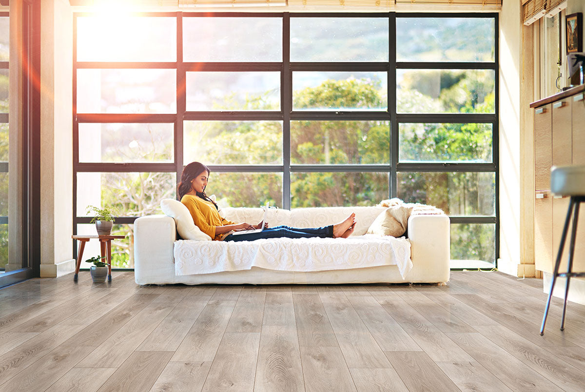 A woman uses a laptop on a white sofa in a bright living room with large windows and MSI XL Cyrus Whitfield Gray Luxury Vinyl Planks (22.44 SQFT). Sunlight streams in, greenery is visible, creating an airy, calm space.