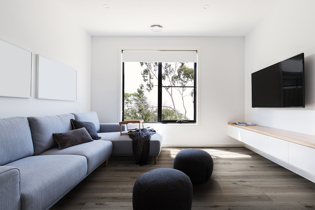 A modern minimalist living room features a light gray sectional, dark cushions, round ottomans, wall-mounted TV, large window with trees outside, and bright MSI XL Cyrus Ludlow Luxury Vinyl Planks flooring for added style.