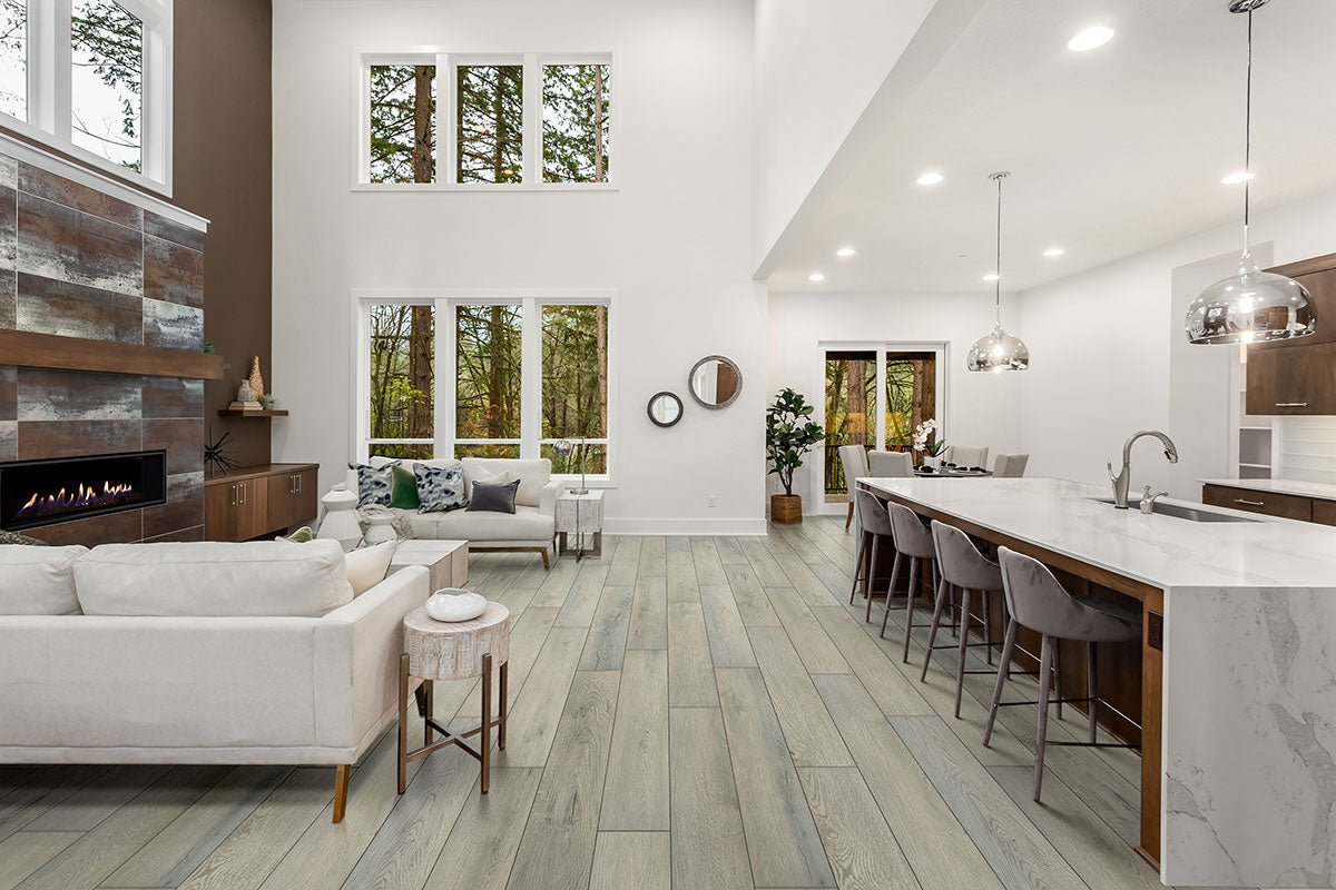 Spacious modern living room and kitchen with tall ceilings, large windows, neutral furniture, MSI XL Cyrus Kardigan Luxury Vinyl Planks flooring (waterproof rigid core), marble island with barstools, pendant lights, and views of trees outside.