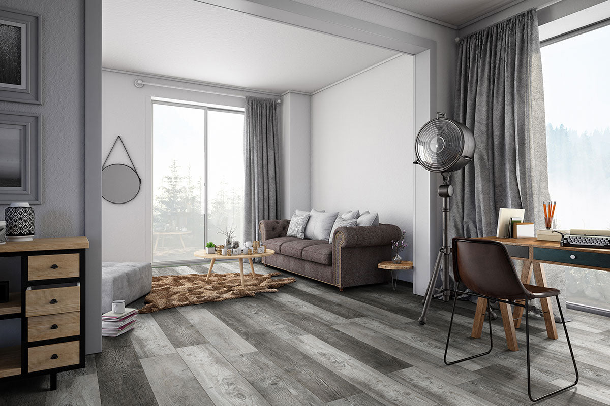 A modern living room features MSI XL Cyrus Boswell Luxury Vinyl Planks (22.44 SQFT/box) waterproof rigid core gray wood flooring, a brown sofa with cushions, shaggy rug, round coffee table, desk with chair, and large windows with sheer curtains.