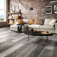 A modern living room featuring a beige sectional, colorful pillows, black oval coffee table, brick accent wall with art, large windows, and wooden shelves is enhanced by MSI XL Cyrus Bracken Hill Luxury Vinyl Planks for stylish, waterproof durability.