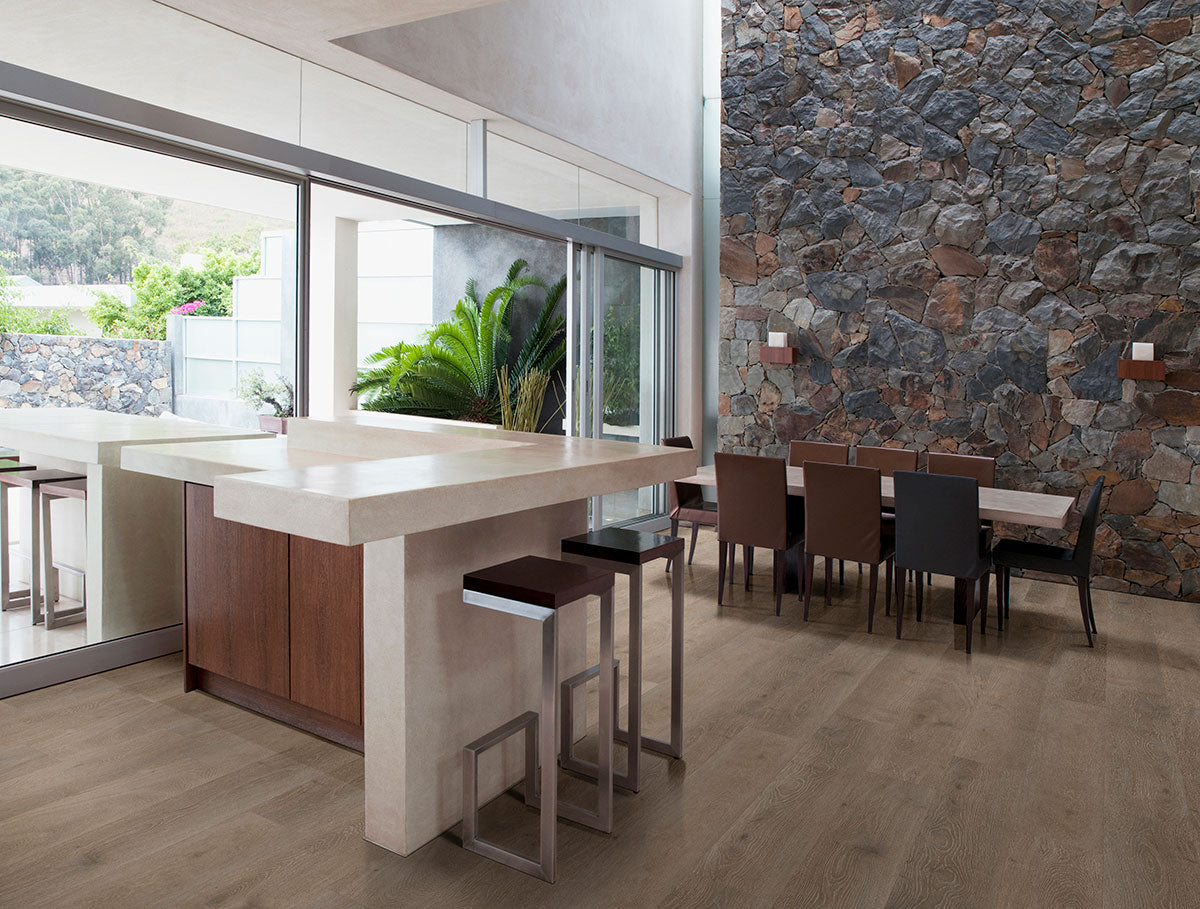 Modern open kitchen and dining area with MSI XL Cyrus Cranton Luxury Vinyl Planks (100% waterproof), a large stone accent wall, white island with bar stools, long dining table, and glass doors overlooking outdoor greenery.