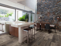 Modern open-plan kitchen and dining area with a large island, MSI XL Cyrus Wolfeboro Luxury Vinyl Planks flooring, bar stools, a wooden dining table with mixed chairs, floor-to-ceiling windows, a stone accent wall, natural light, and greenery outside.