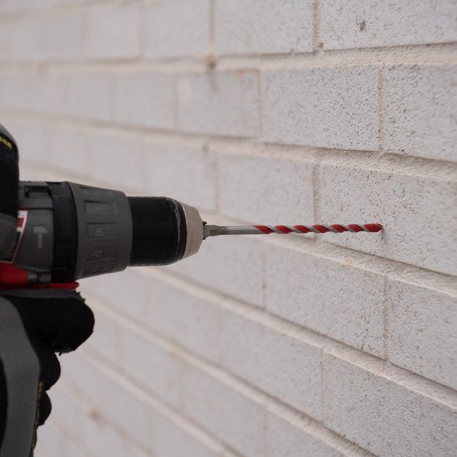 A gloved hand holds a power drill with a Diablo SPEEDemon™ Red Granite Carbide-Tipped Hammer Drill Bit, poised against a white brick wall to create a hole in the mortar, perfect for concrete drilling.