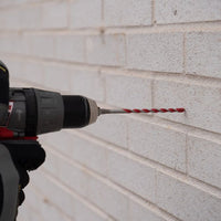 A gloved hand holds a power drill with a Diablo SPEEDemon™ Red Granite Carbide-Tipped Hammer Drill Bit, poised against a white brick wall to create a hole in the mortar, perfect for concrete drilling.
